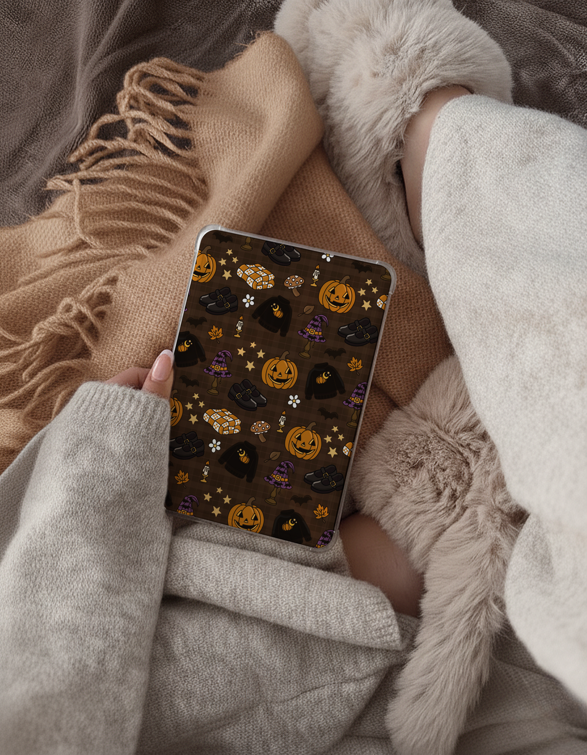 A person holds a smartphone showing a Halloween-themed pattern with pumpkins, ghosts, and stars on a dark brown background, surrounded by cozy beige sweaters, a fringed scarf, and fluffy cream slippers.